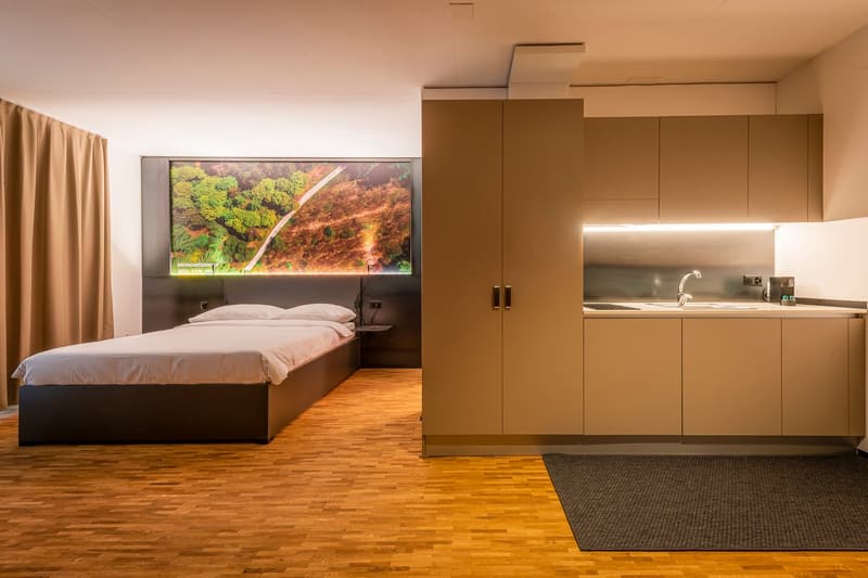 small bedroom with bed, large wall mounted screen, small kitchen with sink and faucet
