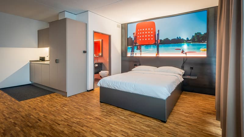 Spacious hotel room with a large bed, wooden flooring, and a flat-screen TV displaying a scenic outdoor view. The room also features a kitchenette area and a bathroom.