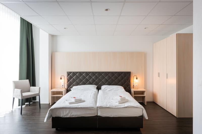 Spacious bedroom with a large bed, bedside tables, and a built-in wardrobe. The room has a modern and minimalist design with white walls, a tufted headboard, and large windows that provide natural light.