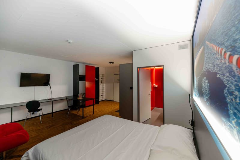 A bedroom with a bed, a desk, a chair, a television, a red chair, and a red wall.