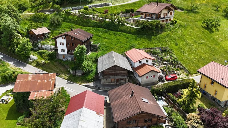 Aerial view of a picturesque village nestled in a lush, green valley. The village features a mix of traditional wooden and stone buildings with red tile roofs, surrounded by well-maintained gardens and greenery. Several houses have balconies or terraces,