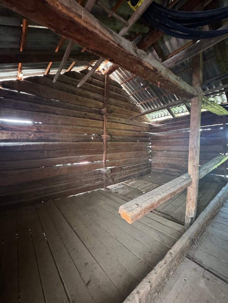 The image shows the interior of an old, wooden attic space. The attic has a slanted roof with exposed wooden beams and rafters. The floor is made of wooden planks, and there are various items and materials stored in the attic.