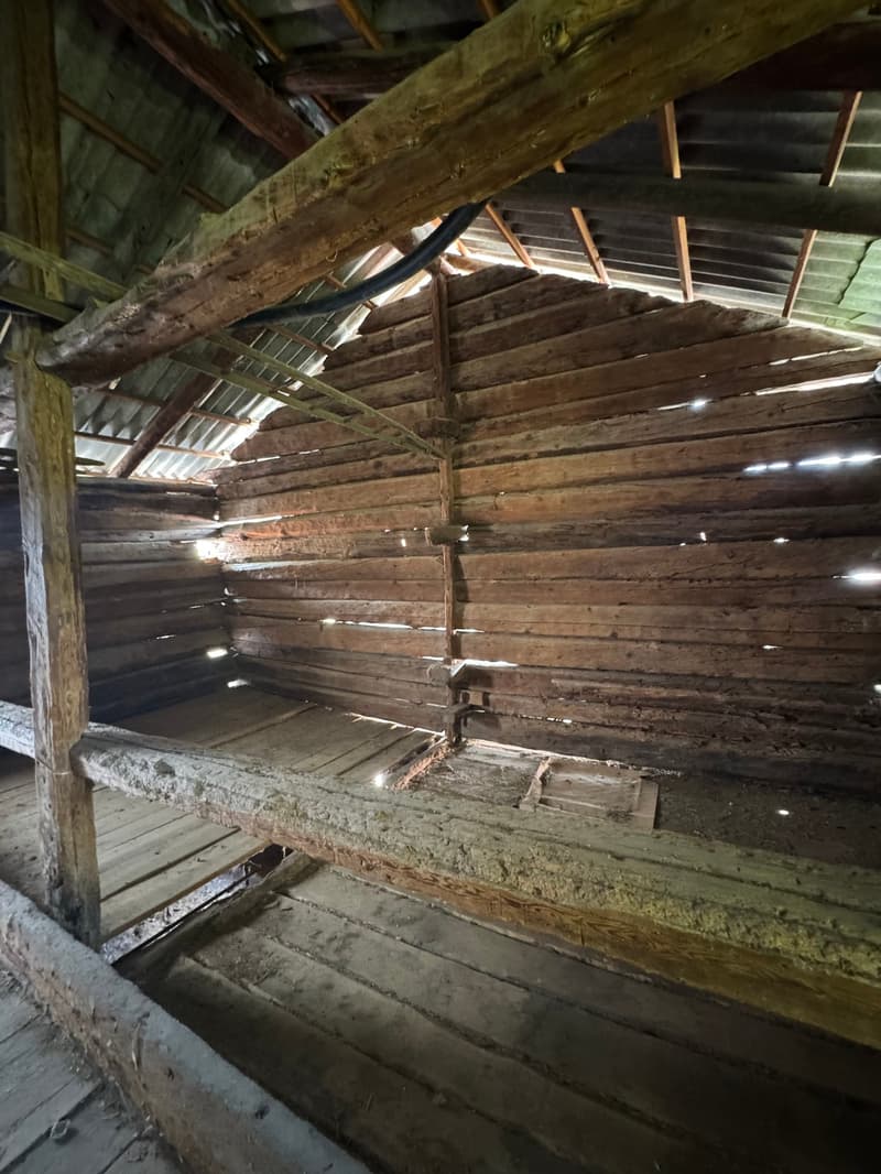 The image shows the interior of an old, wooden barn. The walls are made of rough, weathered wooden planks, and the ceiling is supported by large wooden beams. The floor appears to be made of wooden boards as well. The overall atmosphere is dark and rustic