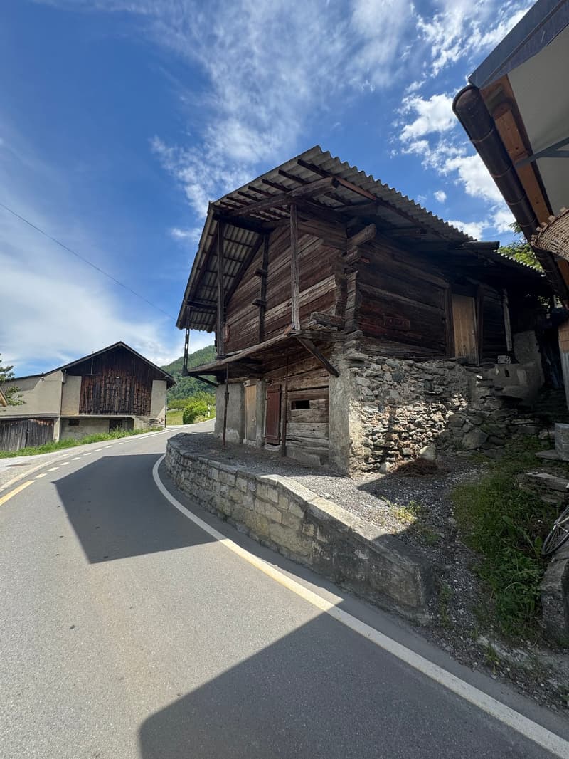 An old, wooden building with a slanted roof and stone foundation, situated along a paved road in a rural, mountainous area. The building appears to be a traditional structure, likely used for storage or agricultural purposes.
