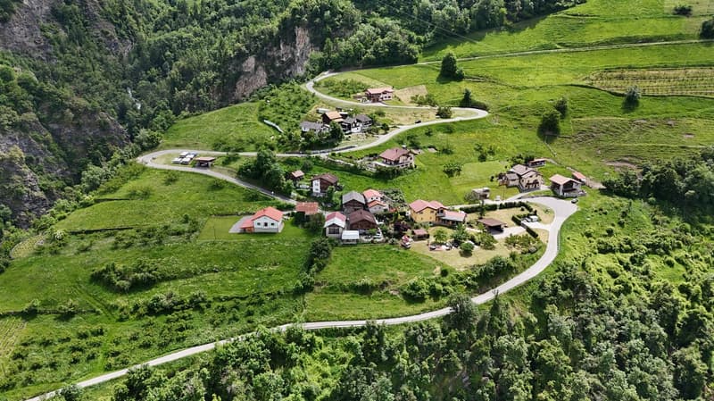 Aerial view of a picturesque mountain village nestled in a lush, green valley. The village features a winding road, several houses and buildings with red roofs, and is surrounded by rolling hills and dense forests.