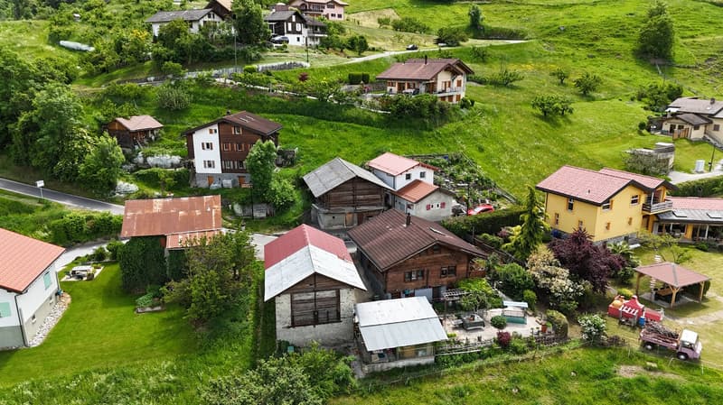 Aerial view of a picturesque village nestled in a lush, green valley. The village consists of traditional Alpine-style houses with red tile roofs, surrounded by well-maintained gardens and orchards. There are several small outbuildings and a few vehicles