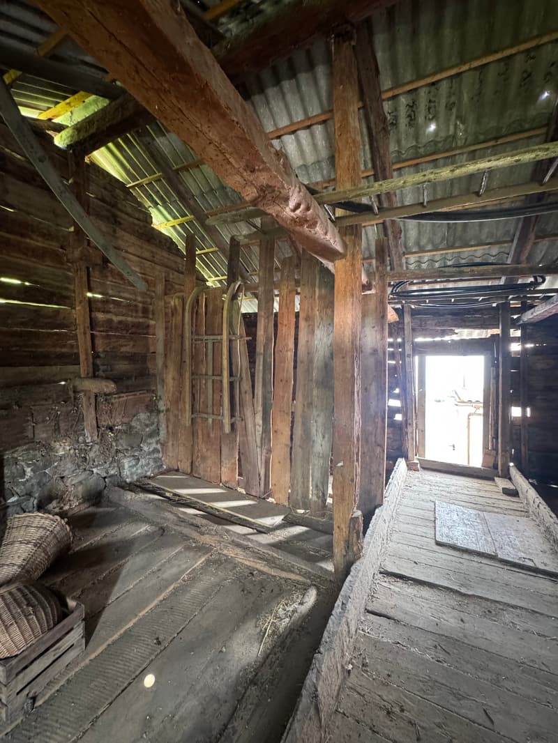 The image shows the interior of an old, dilapidated wooden structure. The walls are made of rough, weathered wooden planks, and the ceiling is supported by large wooden beams. The floor is also made of wooden planks, some of which appear to be damaged or