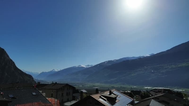 The image shows a scenic mountain landscape with a clear blue sky and a bright sun. In the foreground, there are rooftops of buildings, suggesting a small town or village nestled in the valley. The mountains in the background appear to be snow-capped, cre