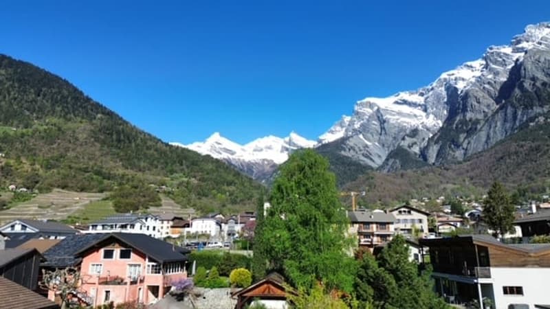 The image shows a scenic mountain landscape with snow-capped peaks in the background. There are several buildings and houses in the foreground, surrounded by lush greenery and trees. The sky is a clear, bright blue, creating a picturesque and serene setti