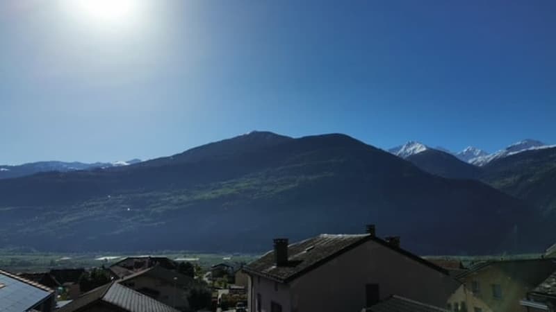The image shows a scenic mountain landscape with a small town or village in the foreground. The mountains in the background appear to be snow-capped, indicating a mountainous region. The sky is clear and blue, and the sun is shining brightly, creating a p