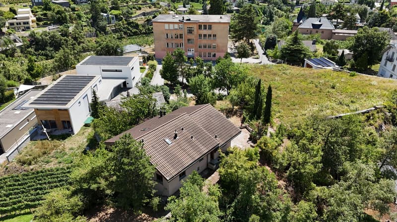 House with brown roof, surrounded by greenery, multiple buildings in the background, solar panels on some roofs, driveway in front of the house, area in the foreground with grass and trees.