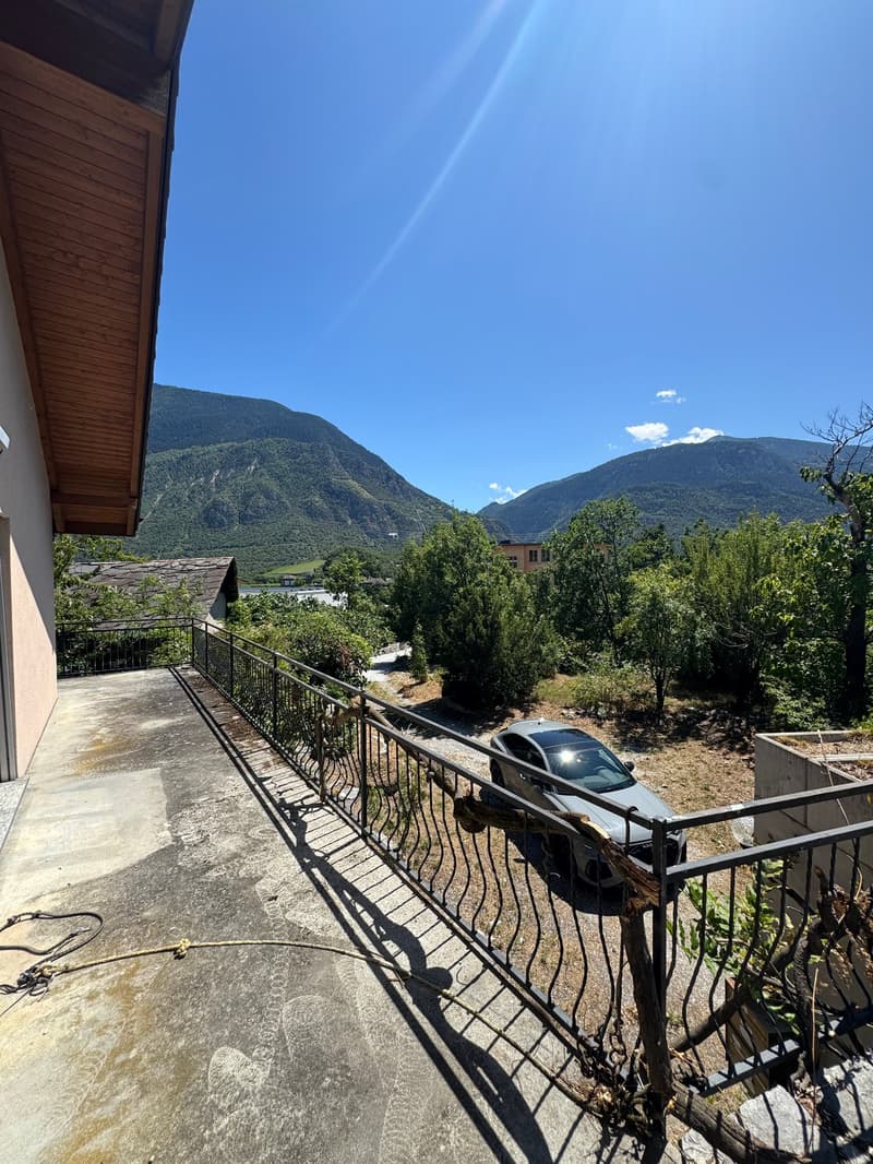 balcony with railing, car parked on driveway, trees, plants, mountains in background, bright clear sky
