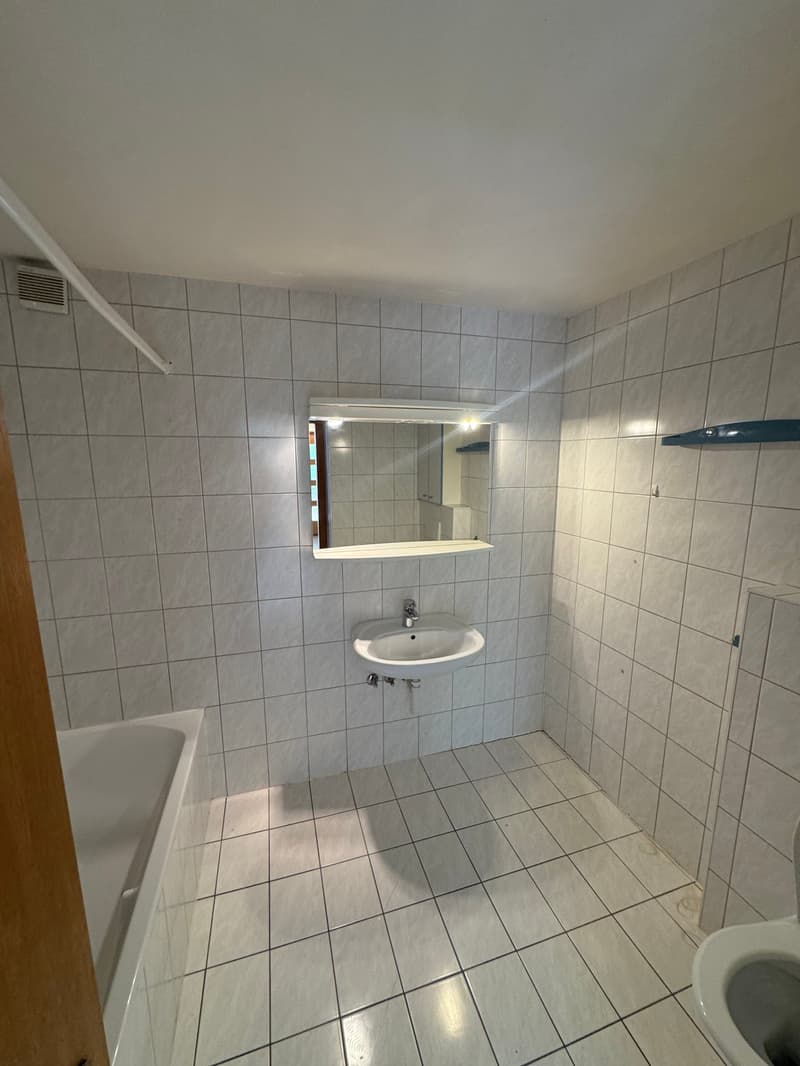 White tiles on walls and floor, white bathtub, white sink, mirror above sink, white toilet, ventilation vent on wall