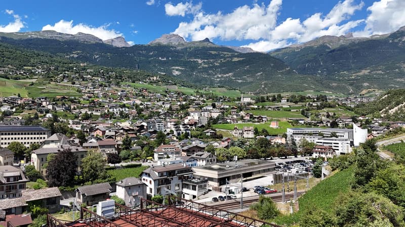 Hilltop village with multiple buildings of various architectural styles, surrounded by trees and greenery, expansive mountain range in the background, visible parking lots and vehicles.