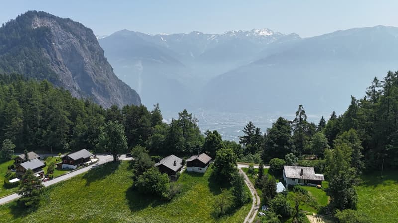 The image shows a scenic mountain landscape with a small village nestled in the valley below. The village consists of several traditional alpine-style houses surrounded by lush green forests and meadows. In the background, towering snow-capped peaks can b