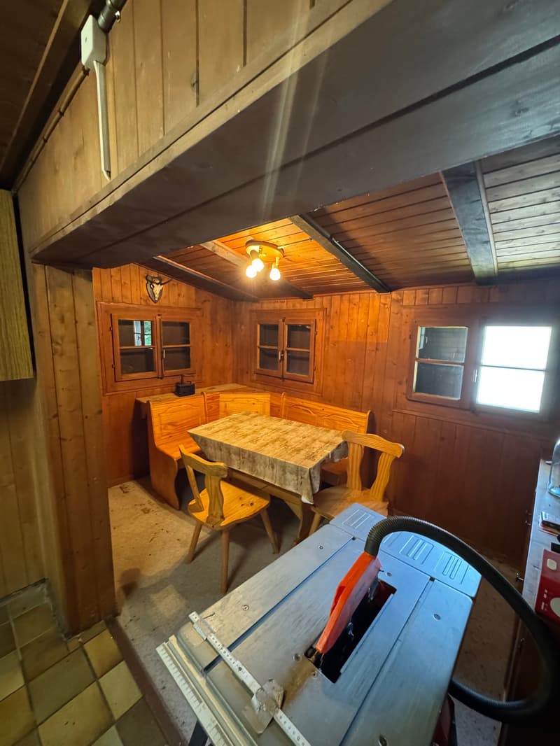 The image shows the interior of a rustic wooden cabin or shed. The walls and ceiling are made of wooden panels, and there is a wooden table with benches in the center of the room. The room is illuminated by a light fixture with multiple bulbs. There are a