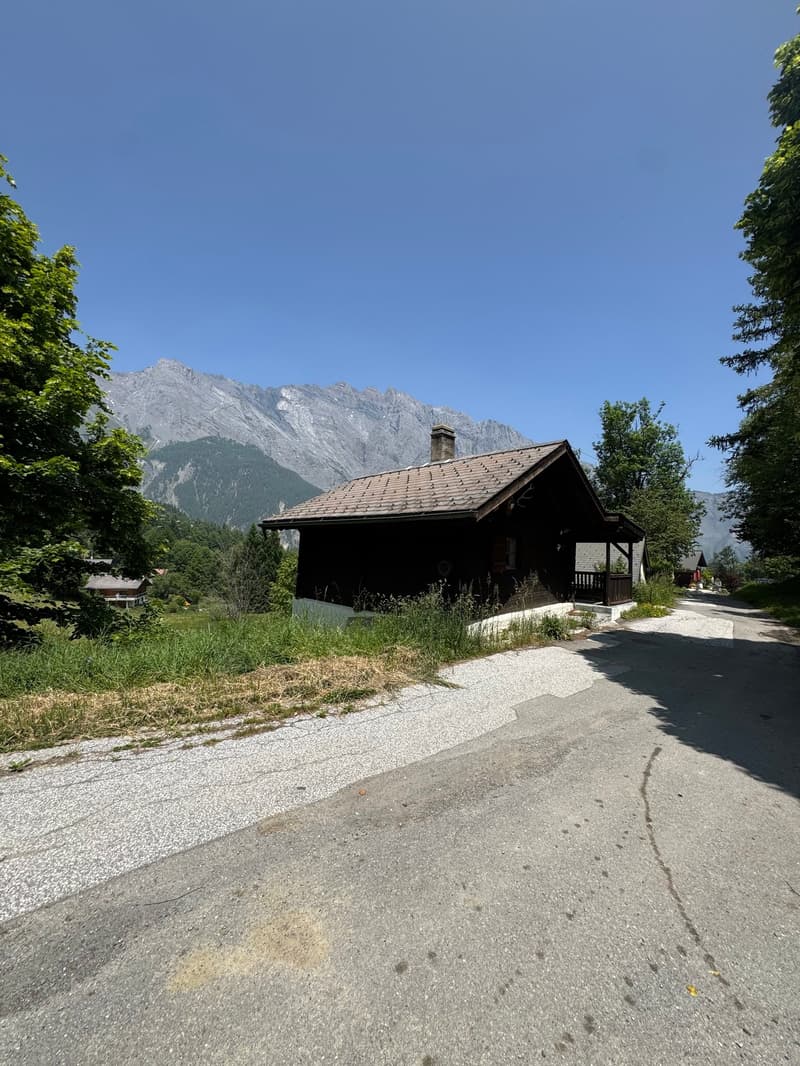 A wooden cabin-style house with a balcony, surrounded by lush greenery and mountains in the background.
