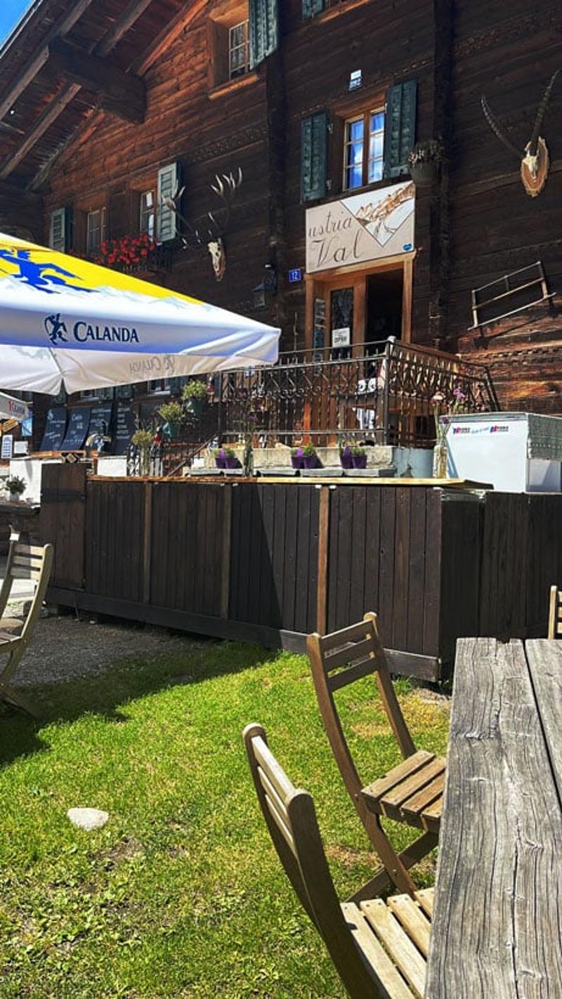 Rustic wooden building with outdoor seating area, umbrellas, and a bar counter. The building has a balcony or terrace overlooking a grassy area.