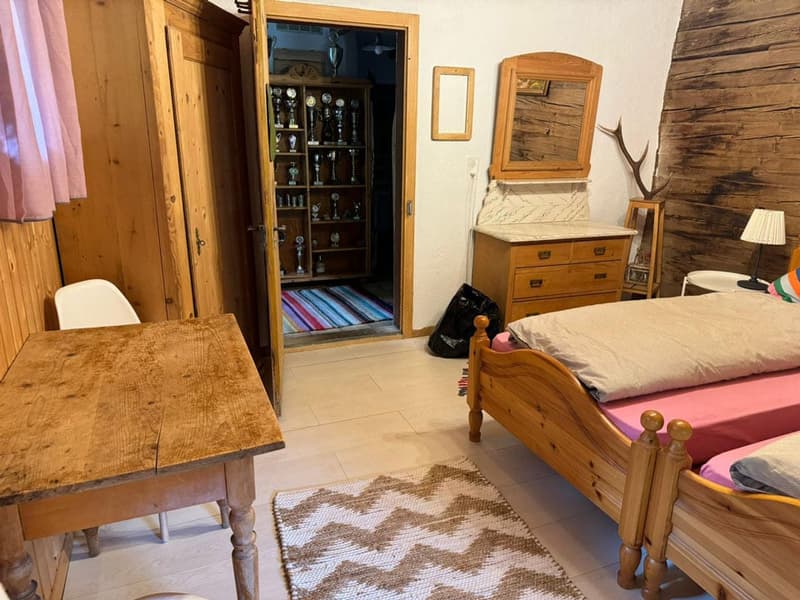 Rustic bedroom with wooden walls and furniture, including a dresser with trophies, a bed, and a table. There is also a fireplace and what appears to be a washing machine visible in the room.