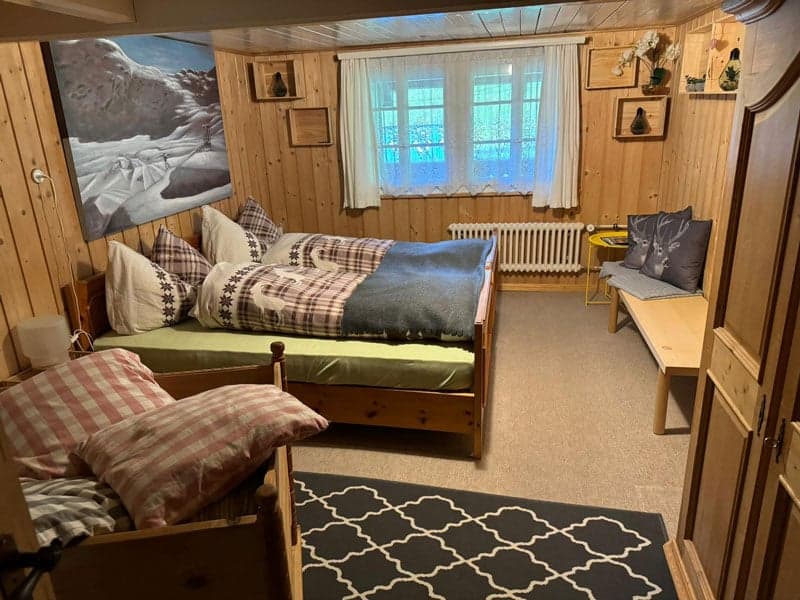Cozy wooden cabin-style bedroom with plaid bedding, large window, artwork on the wall, and a fireplace visible in the corner.