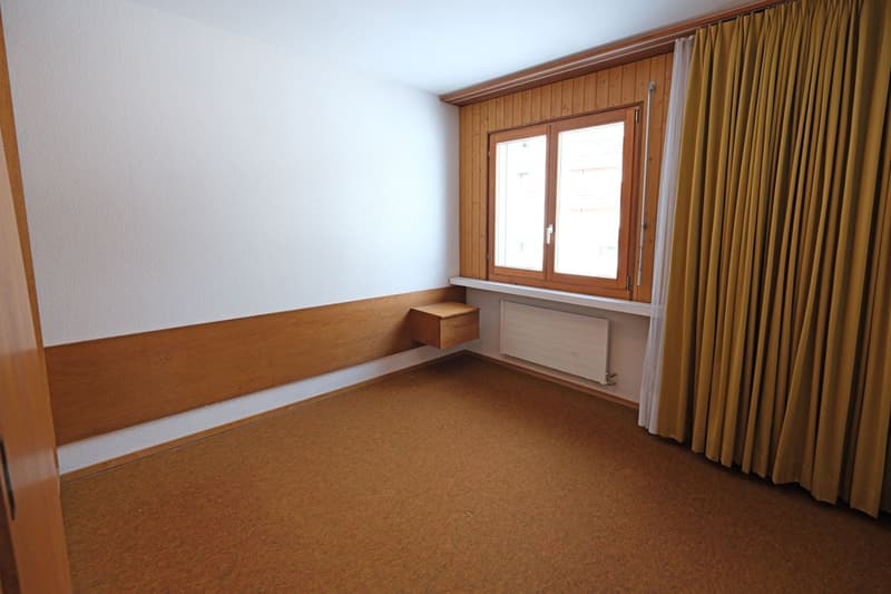 Empty room with white walls, wooden paneling, carpeted floor, a window with a radiator, a wooden storage box.