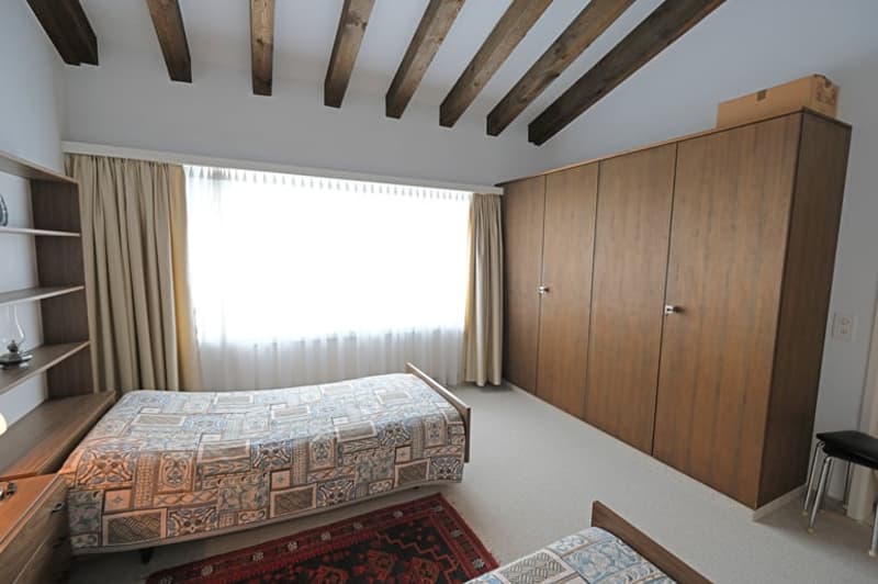 bedroom with two beds, wooden wardrobe, wooden shelves, rug, lamp, window with curtains