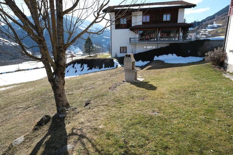 House with balcony, surrounded by snow-covered mountains, grassy field, and a tree