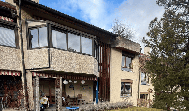 2 story house, beige walls, wooden slats, large balcony, awnings, patio with table and chairs, small garden, clear sky, trees