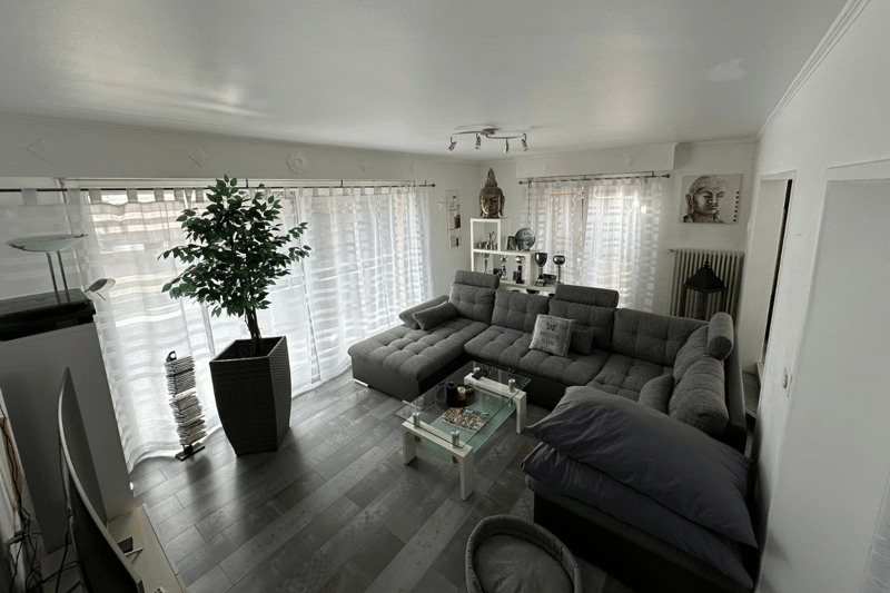 living room with modern grey sofa, glass center table, white curtains, potted plant, decorative items, lamps, radiator, TV stand with flat screen TV