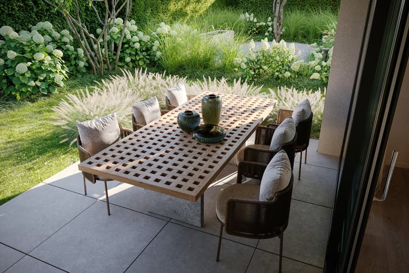 Wooden lattice dining table with cushioned chairs, surrounded by lush greenery including hydrangea bushes and ornamental grasses. The dining area is located on a paved patio with a view of the garden.