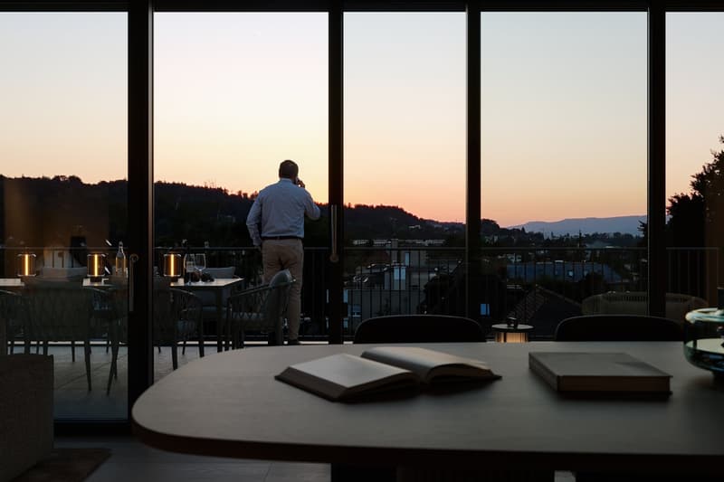The image shows a balcony or terrace overlooking a scenic view at sunset. There is a table with wine glasses and a book on it, suggesting this is a relaxing outdoor space. A person is standing at the railing, taking in the view.