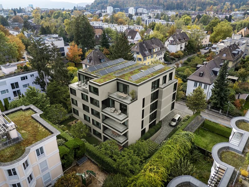 Multi-story apartment building with solar panels on the roof, surrounded by trees and greenery, with balconies and parking spaces visible.