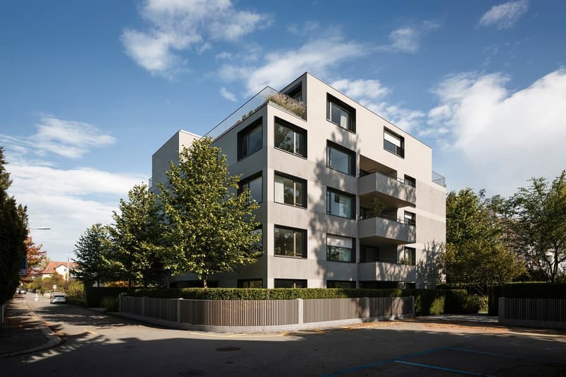4-story modern apartment building with a beige exterior, large windows, and balconies. The building is surrounded by trees and landscaping, with a paved driveway in front.