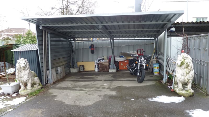 Sheltered carpark with two stone lion statues on both sides, motorcycle, barrels, trolley and buckets.