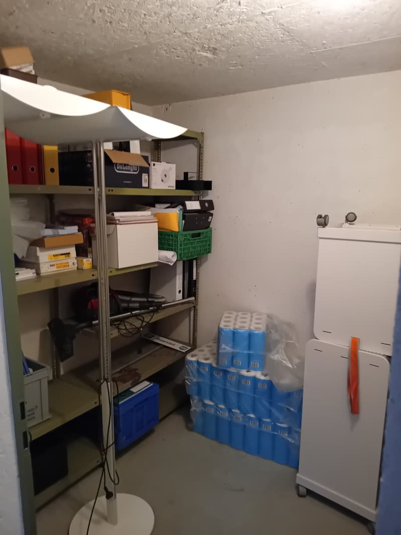 Storage room with shelves, boxes, and a vacuum cleaner. A white refrigerator with an orange handle is on the right side.