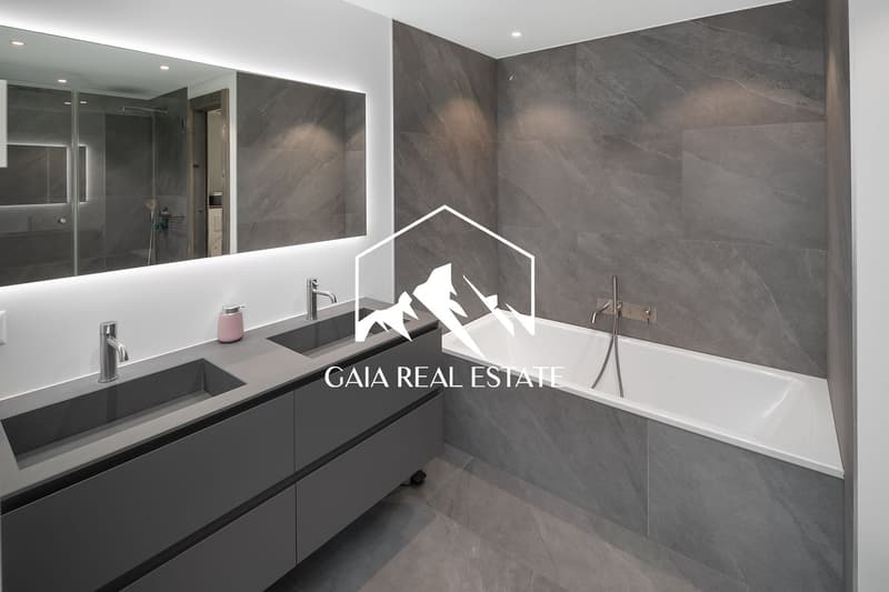 Large bathroom with grey marble-like tiles covering the walls and floor. Double vanity with black countertop and sink, freestanding bathtub, and glass shower enclosure.