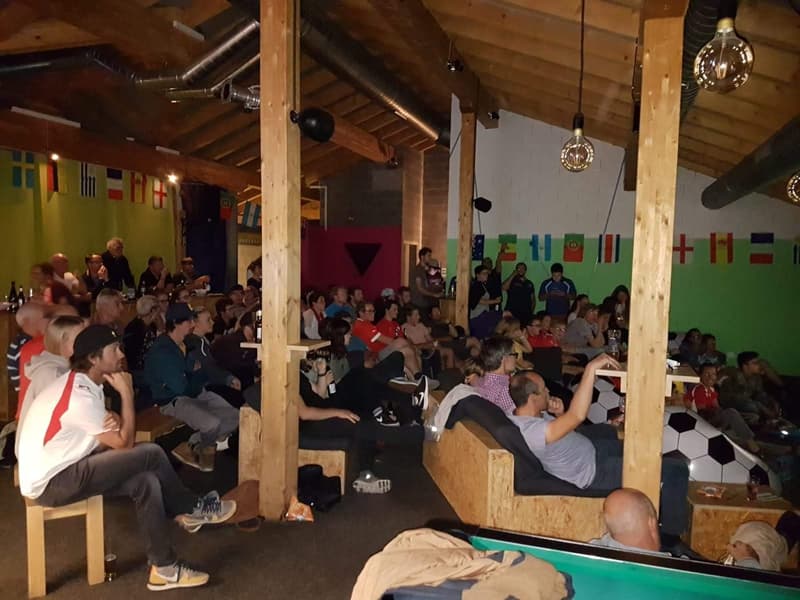 People sitting on benches around a room with wooden columns, flags on the wall, tables with soccer balls and bottles, wooden stools.