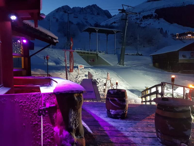 Wooden deck with a few barrels on it, the deck has purple lights on it, wooden posts with lights and the background has mountains covered in snow.