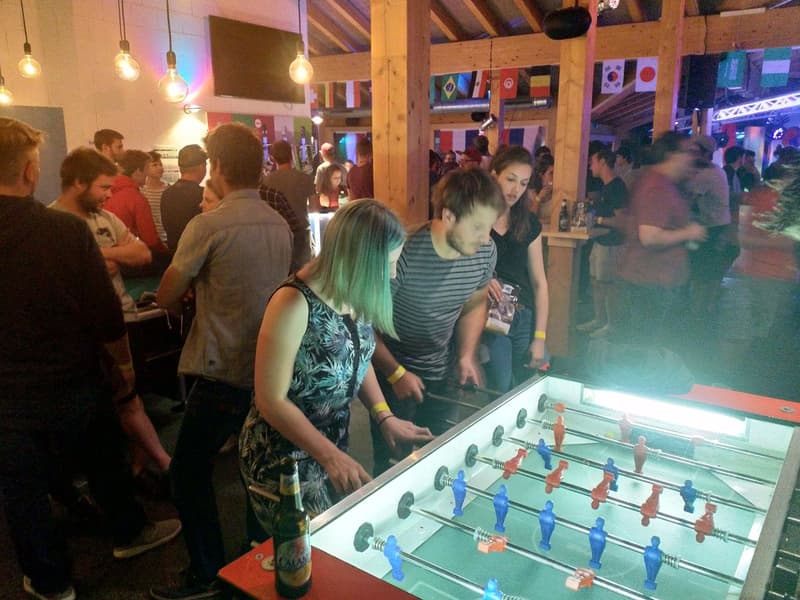 Foosball game with people in crowded area, tables with other drinks.