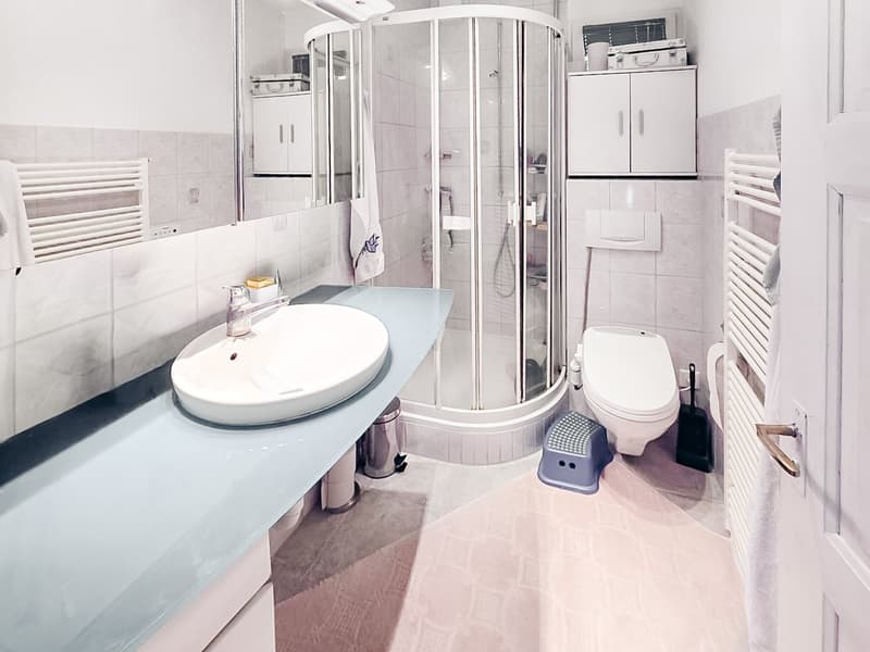modern bathroom with white tiles, glass shower enclosure, white sink, white toilet, white cabinets, white towel radiators, trash can, and a step stool