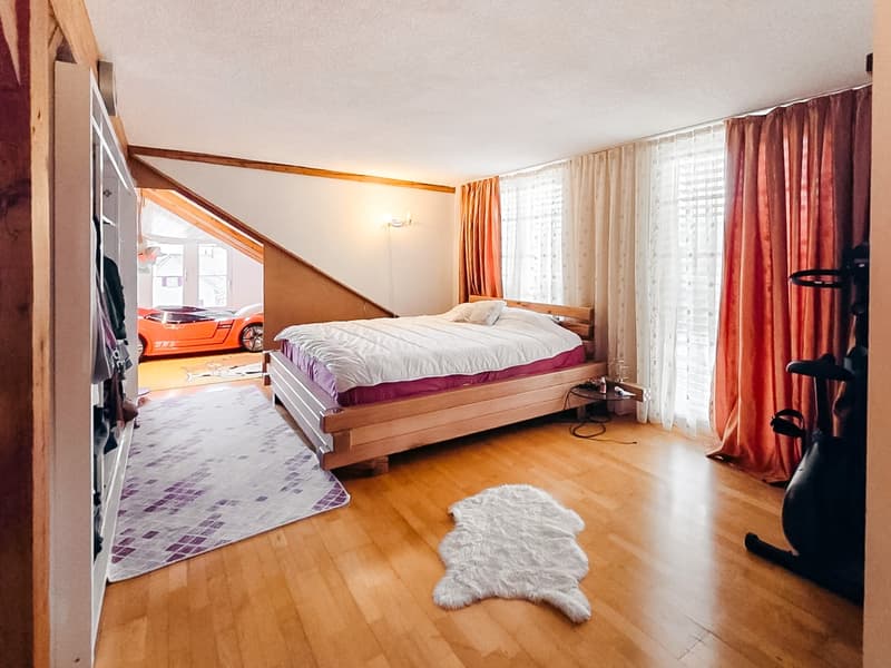 Bedroom with wooden bed, white rug, wooden floor, white walls, large windows, exercise bike, car visible in the background