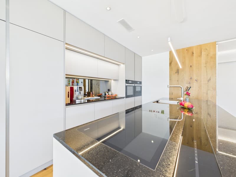 Modern kitchen with granite countertops, sleek white cabinetry, induction hob, stainless steel appliances, a wine rack, a sink with a stylish faucet, wooden elements, pendant lights, a refrigerator, and a countertop with fruits.