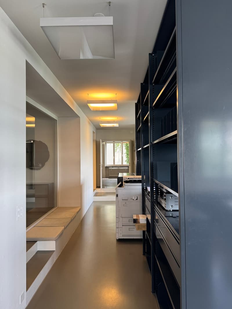 Corridor with white walls, gray floor, large windows, a bench, and a shelf with various items