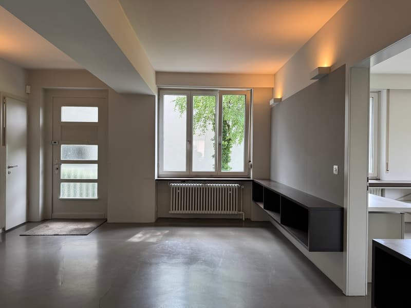 Empty room with white walls, gray floor, white door, large window, radiator, and a rug.