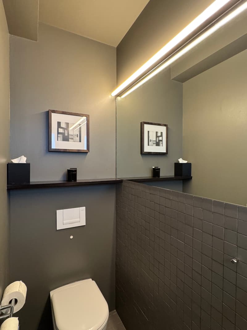 small bathroom with gray walls, white toilet, white toilet paper holder, black shelves with tissue boxes, two framed pictures on the wall, mirror