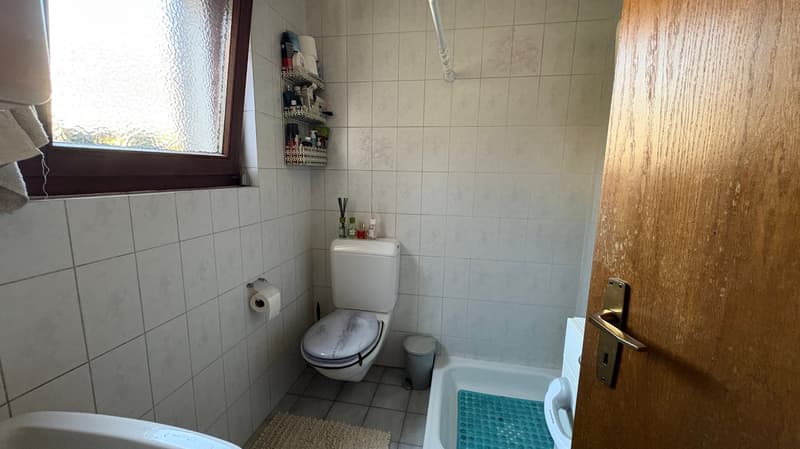 Tiled bathroom with toilet, sink, and shelving unit. There is a washing machine visible in the room.
