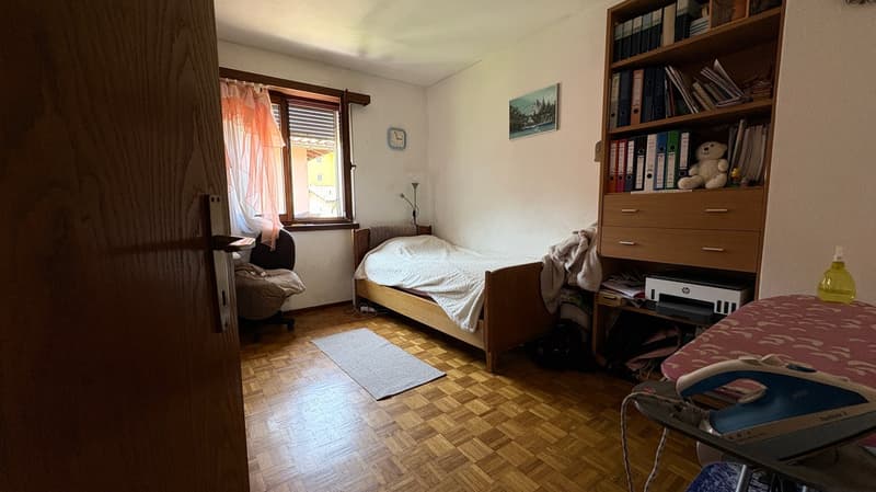 Bedroom with wooden bed frame, bookshelf, desk, and printer. Curtains on the window and a rug on the parquet floor.