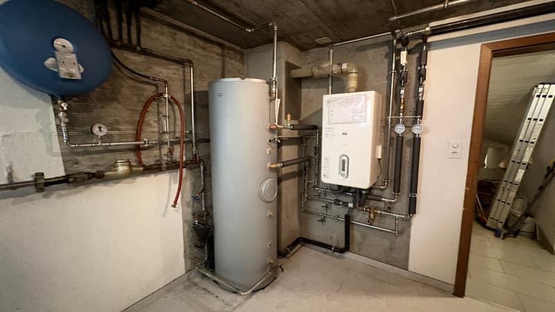 The image shows a utility room with a water heater, various pipes and plumbing equipment, and a washing machine. The room has a concrete floor and exposed wooden beams on the ceiling, giving it a rustic, industrial feel.