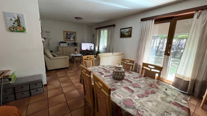 Spacious living room with tile flooring, large windows, sofa, coffee table, TV stand, and dining table with chairs. The room has a cozy and homely feel with various decorative elements and artwork on the walls.