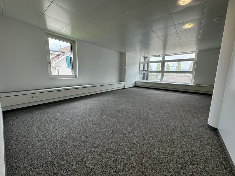 Large open office space with gray carpeted floor, white walls, and recessed lighting. The room has several windows providing natural light.